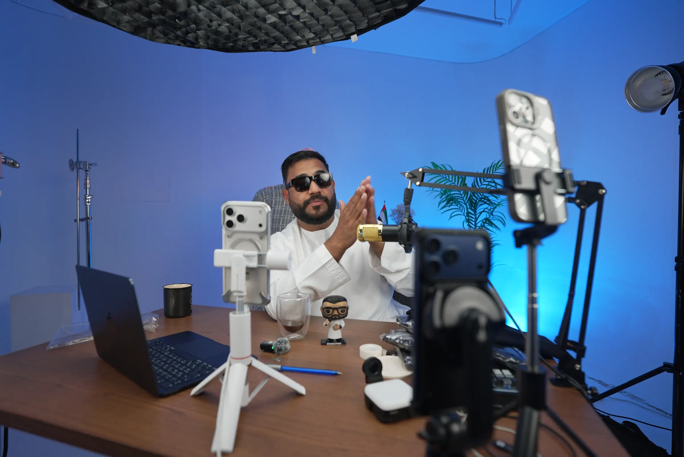 Presenter filming at a desk surrounded by phone rigs, lights and microphones